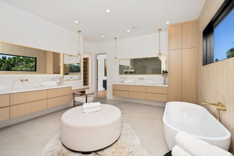 Interior de baño moderno y espacioso con bañera exenta, lavabo doble y espejos grandes. La luz natural que entra por las ventanas ilumina el espacio.