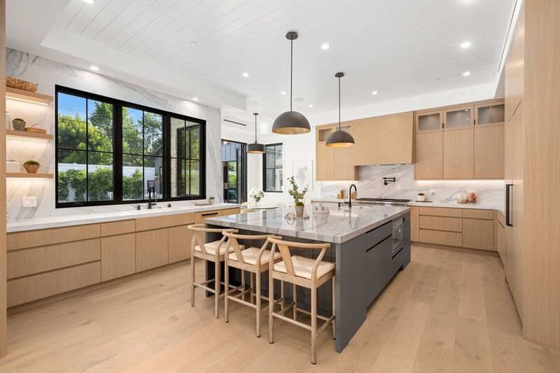 Interior de cocina moderna con una gran isla, gabinetes de madera y luces colgantes, con una gran ventana hacía el jardín.